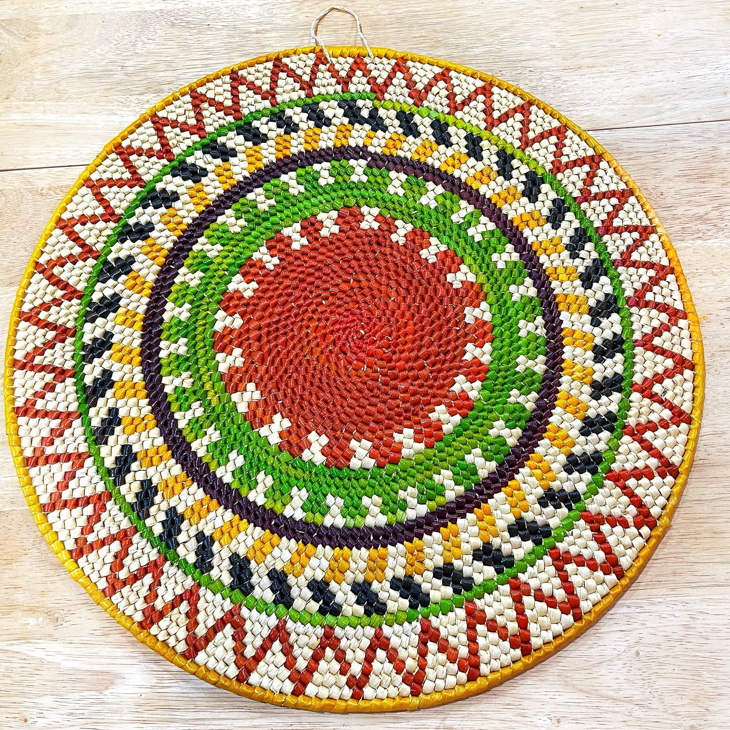 Handwoven Tucumã palm mandala wall art from the Amazon. Sunburst pattern features shades of green, red, yellow, orange, blue, purple, and cream. Symbolizes positive energy and new beginnings. Made by artisans using traditional techniques.