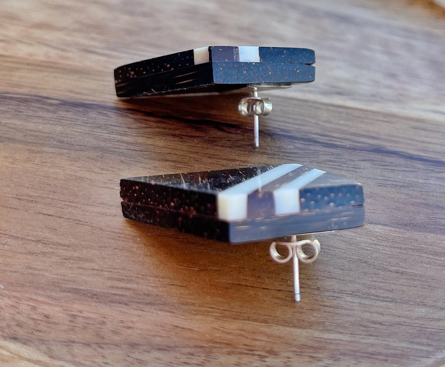 The image shows a pair of stud earrings with a unique design. They are handcrafted from dark, patterned bone with white inlay stripes, showcasing the natural texture of the material.