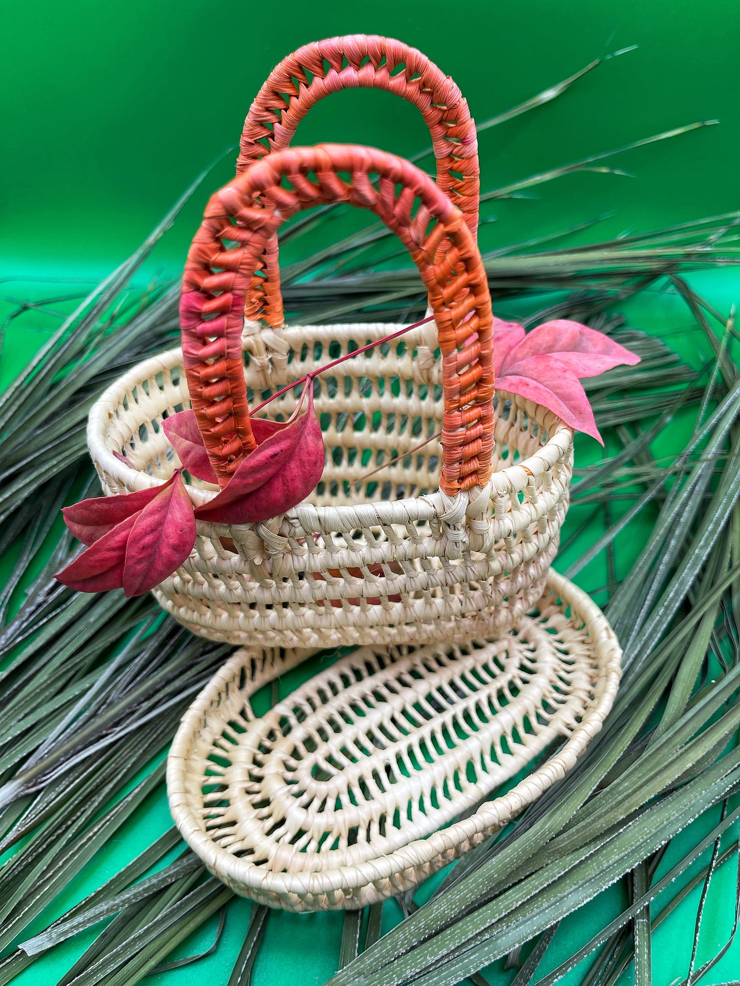 Handwoven mini tucuma basket with vibrant red handles, adorned with pink leaves against a green background with palm sprigs.