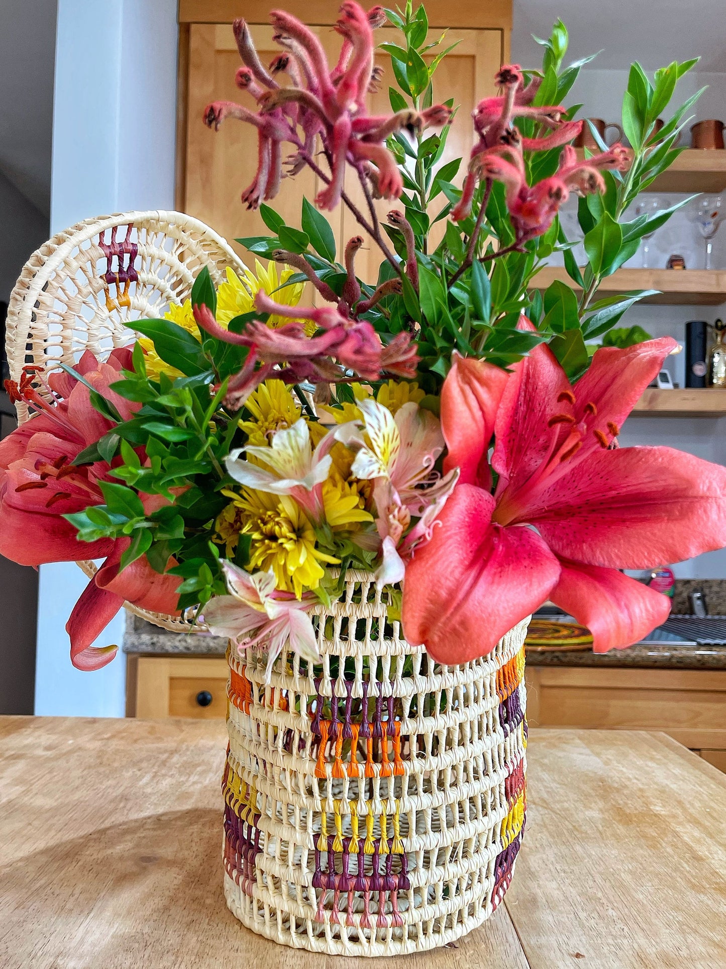 A vivid bouquet springs from a handwoven basket with colorful stripes. Blossoms in pink, yellow, and green hues contrast with the basket's earthy tones, set in a cozy kitchen.