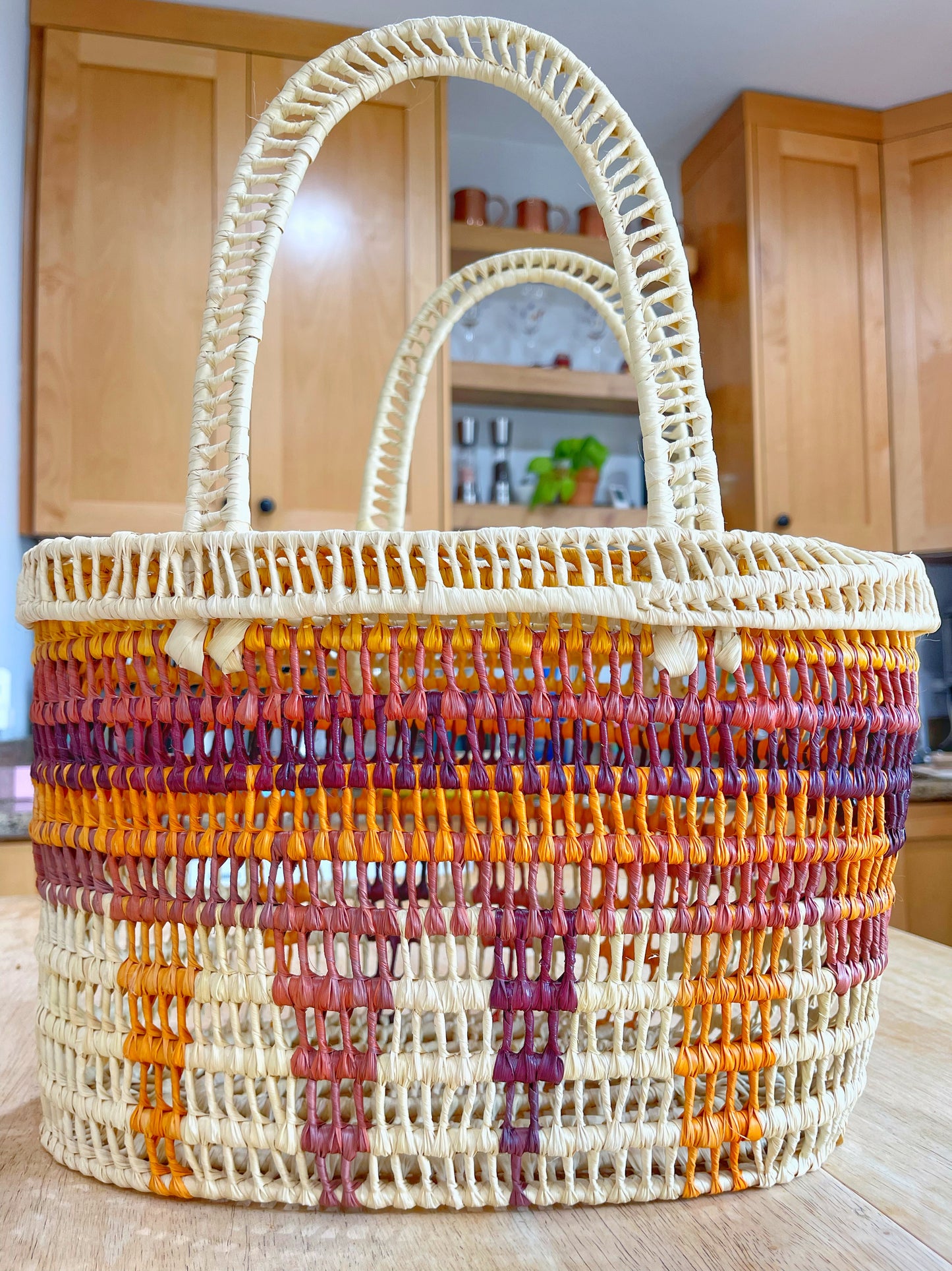 A handwoven basket with a cream base and vibrant stripes of orange, yellow, and burgundy stands out in a warm kitchen setting.