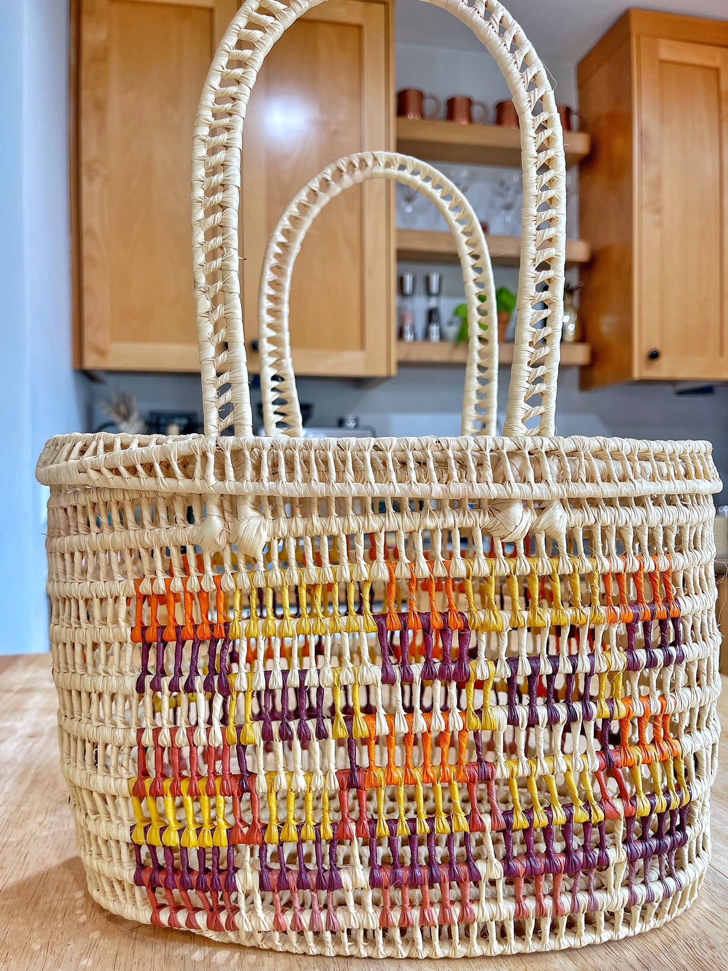 A handcrafted beige basket flaunts a colorful striped center with yellows, oranges, reds, and purples. Its tight weave and sturdy handles are set against a modern kitchen backdrop, showcasing traditional artisanship.