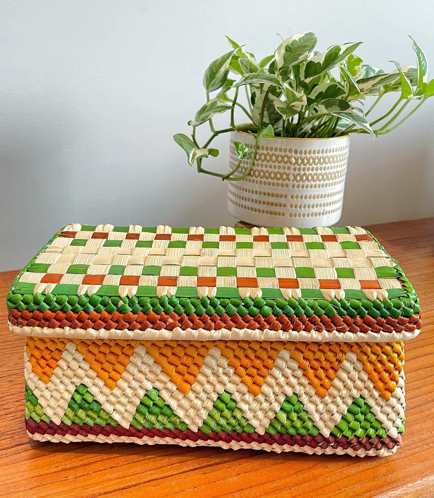Vibrantly handwoven tucuma palm box in a geometric pattern of green, orange, and natural hues, with a green-lined lid, against a homey backdrop