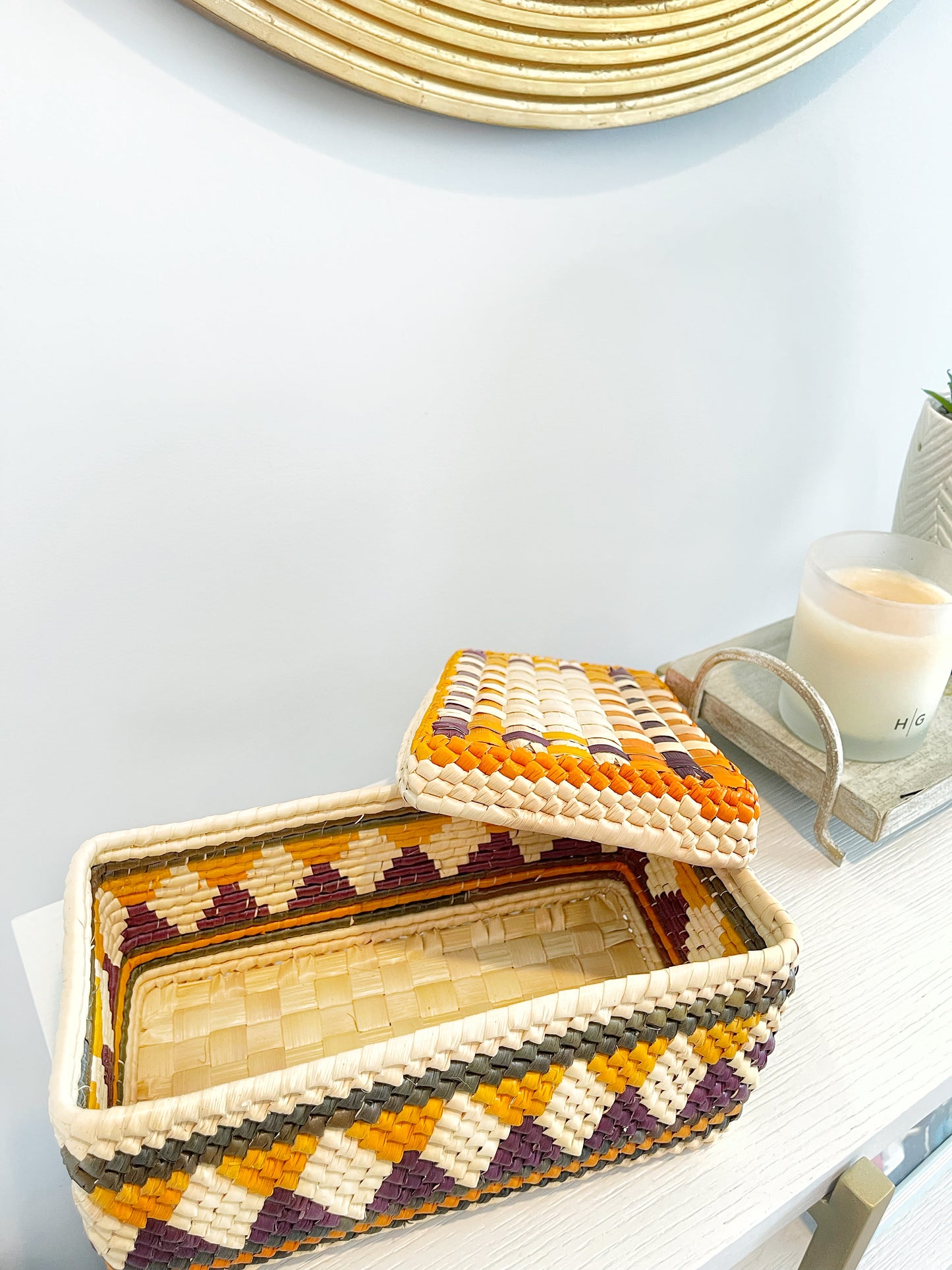 Open handwoven tucuma palm box with intricate patterns in purple, orange, yellow, and black, revealing a light interior, placed on a chic, modern shelf.