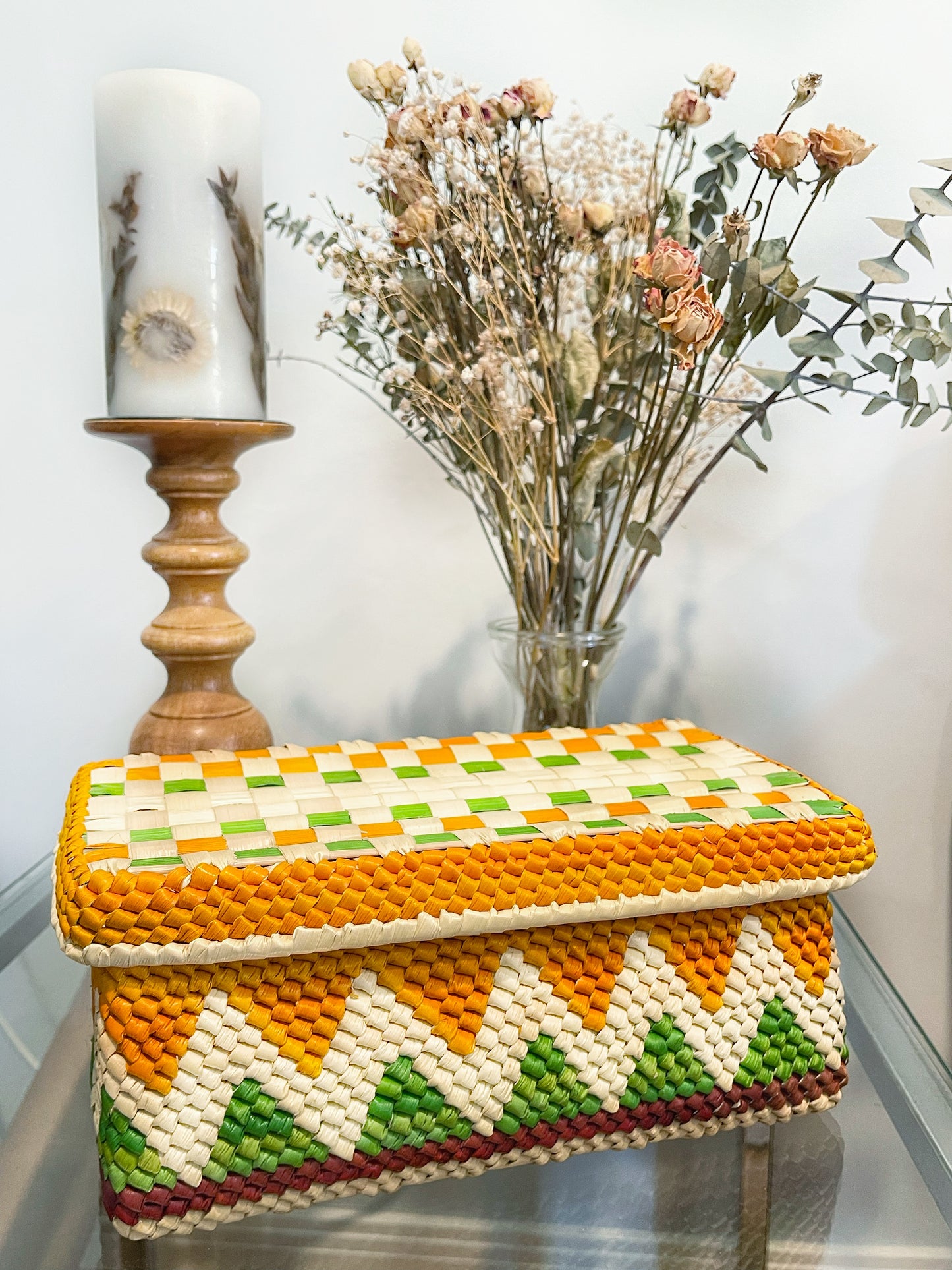 Elegant woven box with a lively pattern of green, orange, and white, evoking traditional Amazonian craftsmanship, accompanied by a dried flower arrangement and a decorative candle.