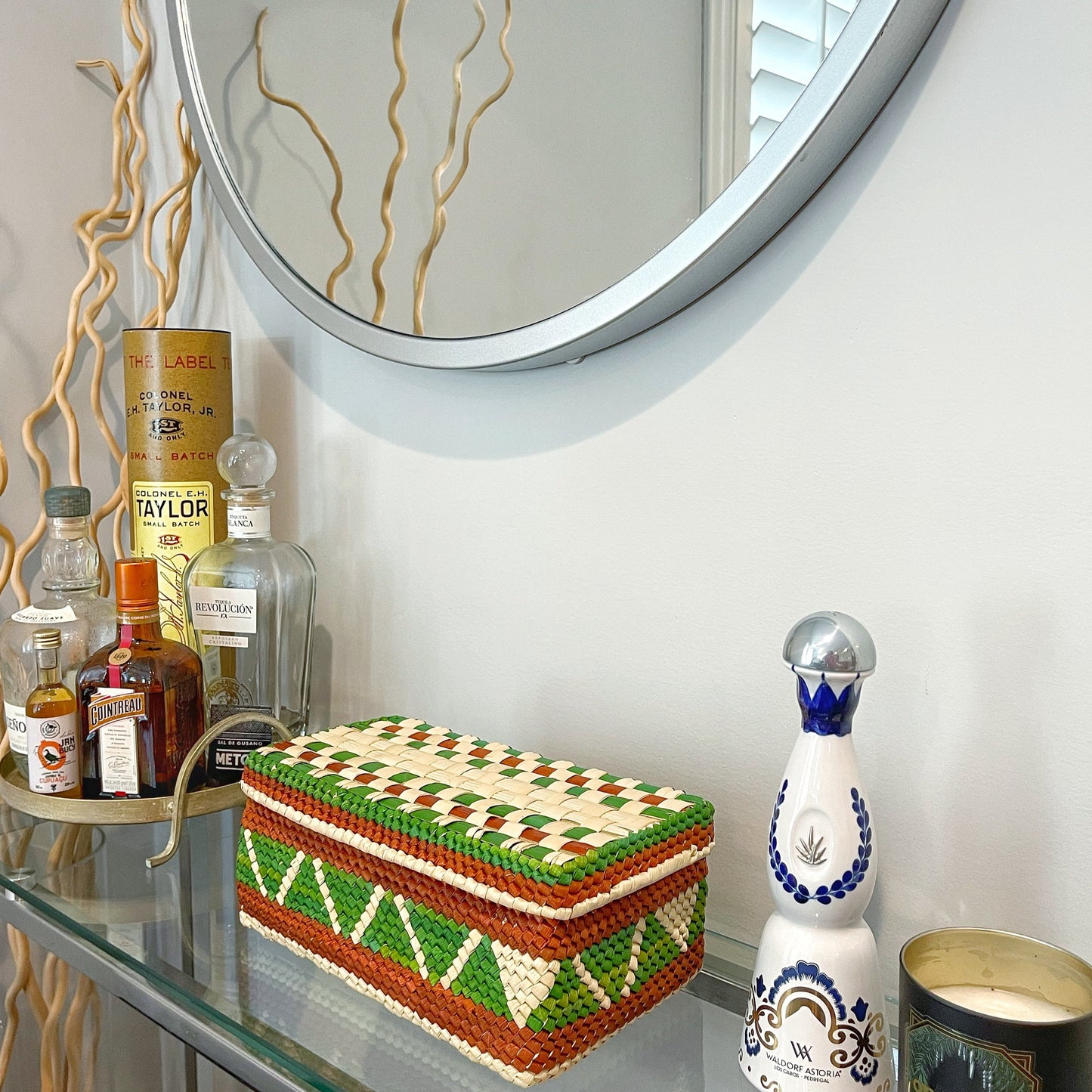 Artisanal tucuma palm box in green, brown, and cream, beside liquor bottles and a decorative blue-topped porcelain flask, on a glass shelf with a round mirror above