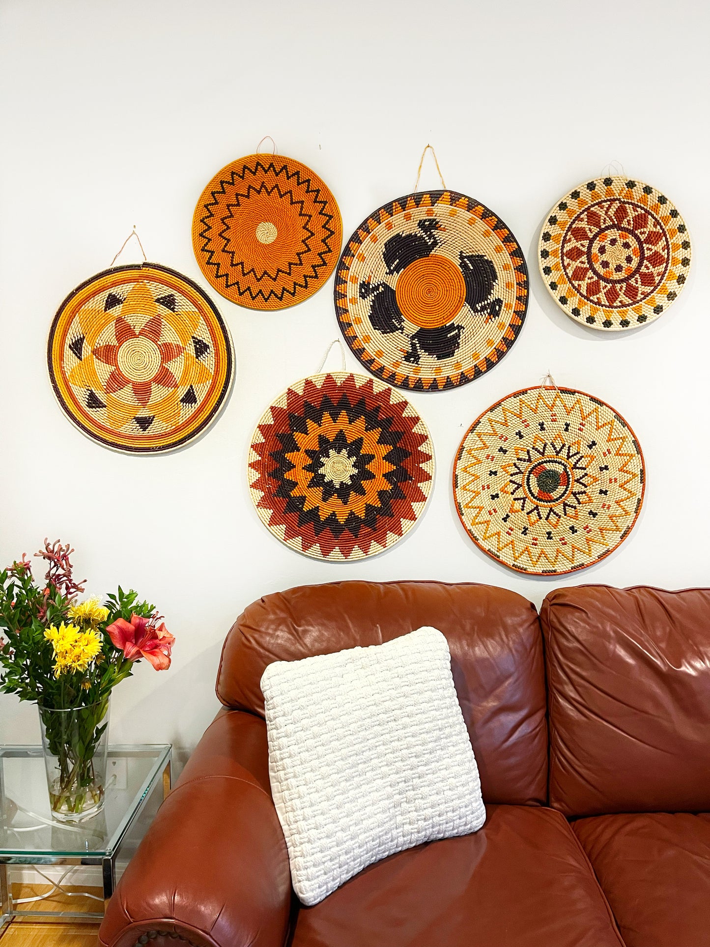 A cozy room with a leather couch, adorned with a white cushion, and a wall above decorated with colorful mandala wall art, plus a glass vase of bright flowers.