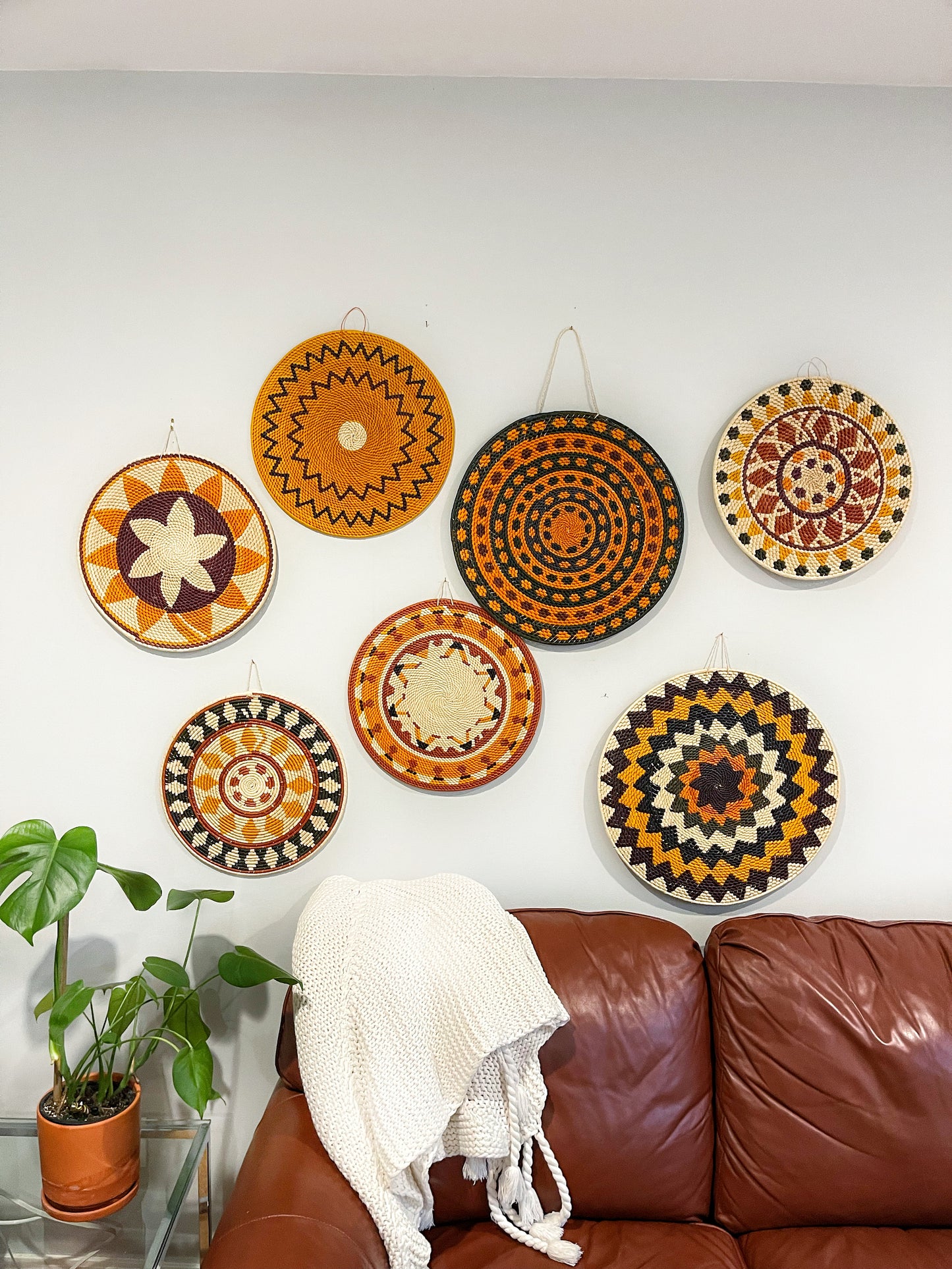 Warm-toned mandalas create a striking focal point above a brown leather couch. A potted plant and white throw add a bohemian touch. Circular designs pop, suggesting an ethnic-inspired decor style.
