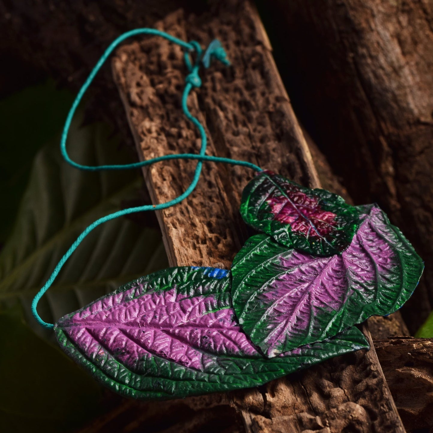 Leaf-shaped necklace, handcrafted from natural rubber blended with açaí seed fibers. The leaf features a realistic design with vibrant green at the edges transitioning to a pink center. A thin turquoise cord suspends the necklace. It's displayed against a background of weathered wood with a visible grain texture.