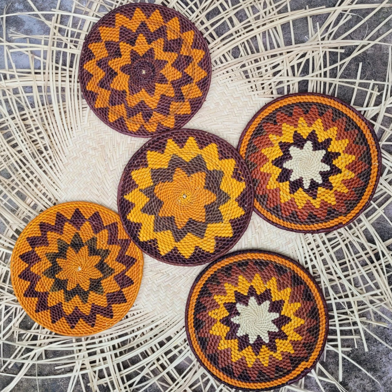 Five circular handwoven trivets on a wicker mat, with starburst patterns in shades of yellow, orange, and dark purple.