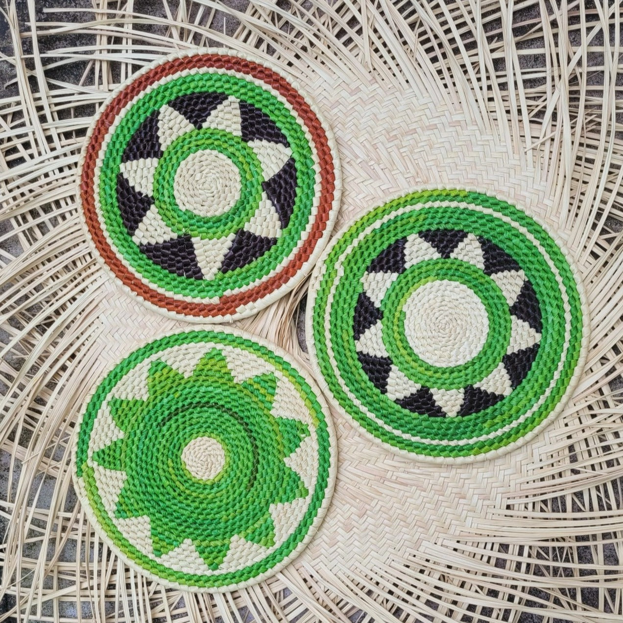 Three handwoven trivets with geometric patterns, made from tucuma palm, in green, white, black, and brown, on a wicker surface.
