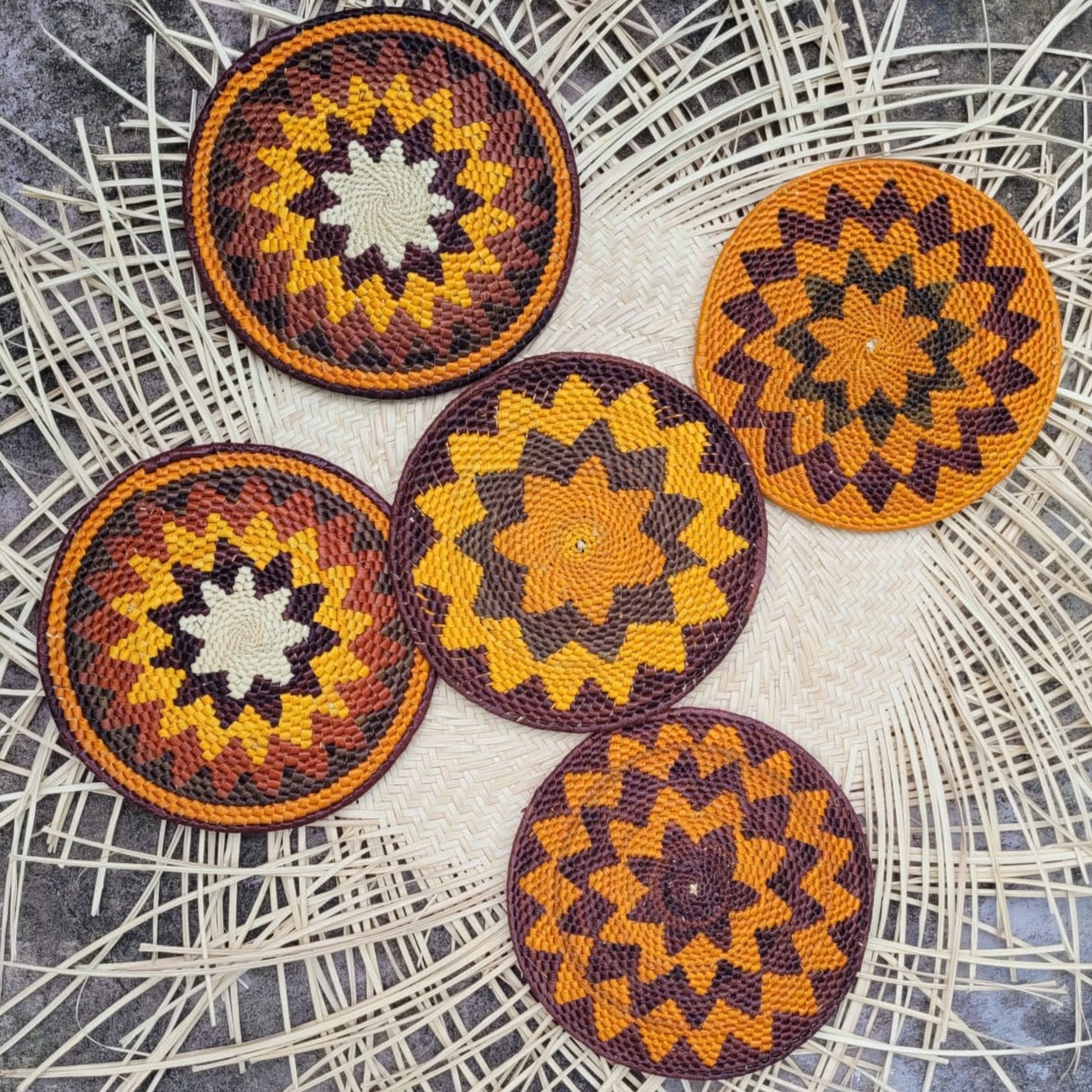 Five round woven trivets on a herringbone mat with starburst patterns in orange, yellow, and dark brown. The intricate designs and colors evoke a warm, natural aesthetic.