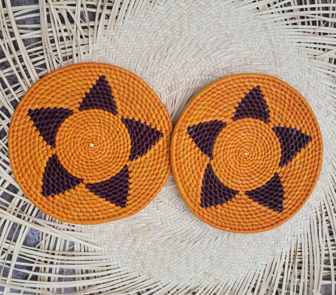 The image displays two handwoven trivets in vibrant orange, featuring a central circular pattern with dark purple triangular motifs radiating outward, resembling stars or flowers.