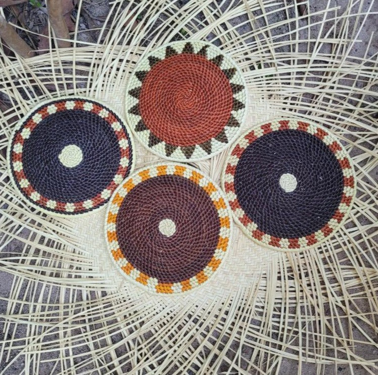 Four circular handwoven trivets, in navy and brown with cream and orange accents, on a wicker mat with a radiating pattern.