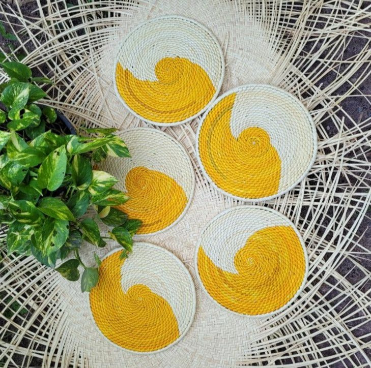 Five cream-colored woven trivets with vibrant yellow swirl patterns displayed on a herringbone-patterned rattan backdrop, complemented by a potted green plant with glossy leaves.