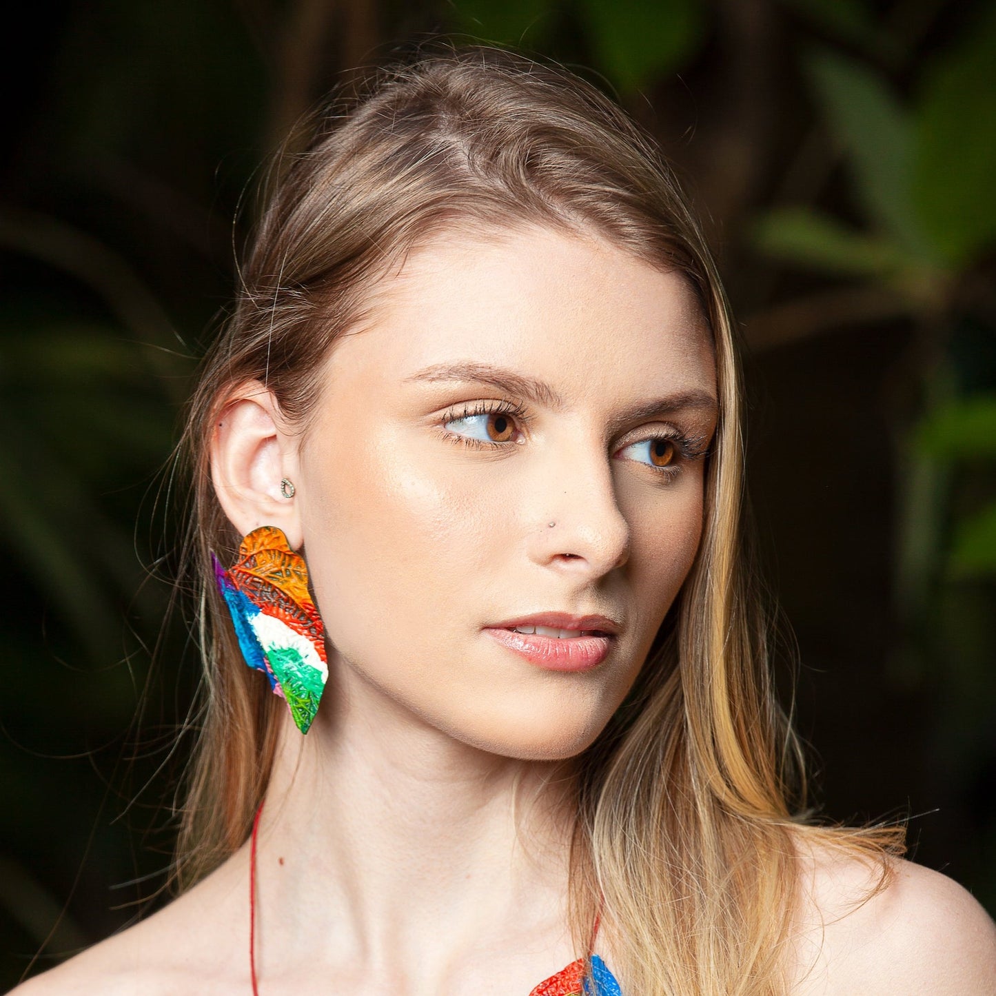 A model wears a leaf-shaped earring, handmade from natural rubber with açaí fiber, nickel-free. It has vibrant orange, green, and blue patterns.