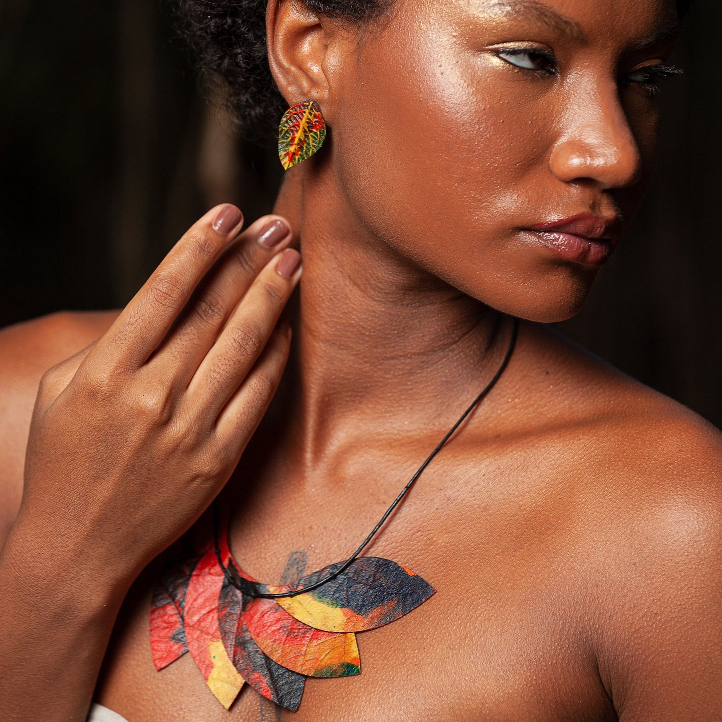 A close-up image of a woman adorned with handcrafted jewelry. She wears an eye-catching necklace made of multiple natural rubber leaves, artfully overlapping in a cascade of autumn hues—crimson, black, and golden yellow. A single, intricately patterned earring is visible, resembling a vibrant leaf crafted from natural rubber and adorned with specks of red, yellow, and green, mimicking the colors of a flame tree.