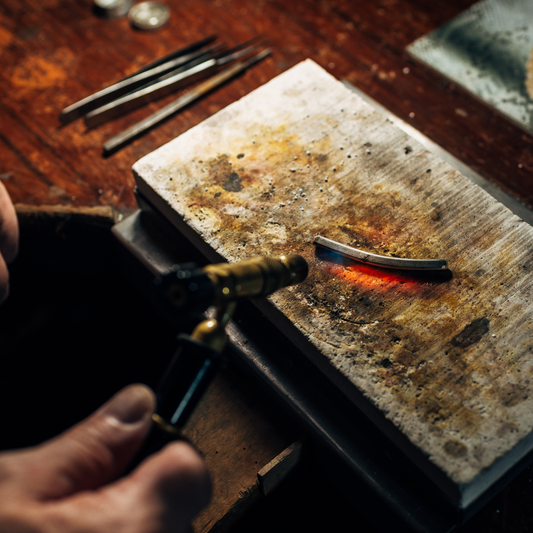 An image of a jewelry-making process with a blowtorch applying heat to a metal piece on a workbench, highlighting the detailed craftsmanship in jewelry design.