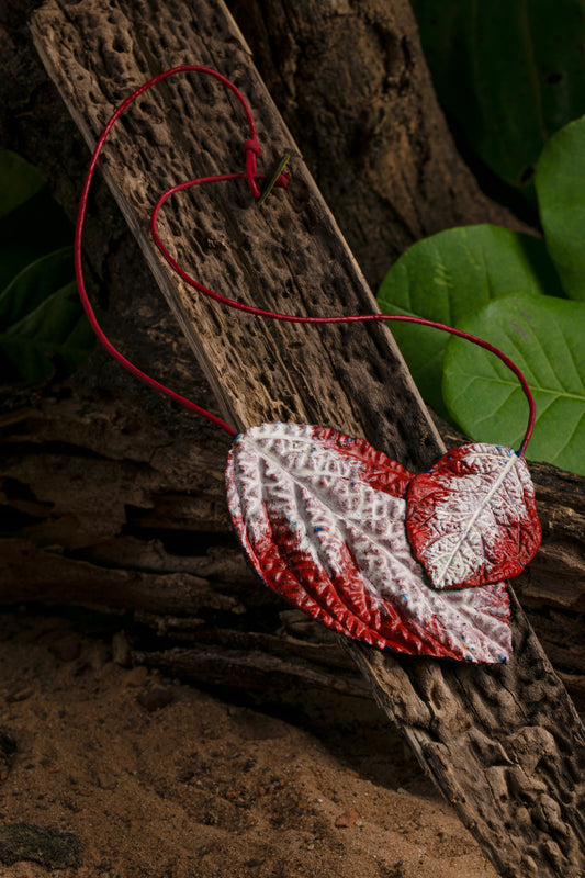 A handmade necklace with two heart-shaped leaves painted in red and white, resting against a rustic wooden backdrop surrounded by green foliage.