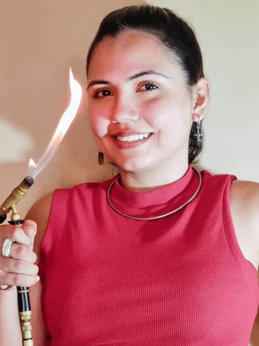 A smiling woman in a red sleeveless top holding a lit blowtorch, showcasing a flame,  during a jewelry-making process.