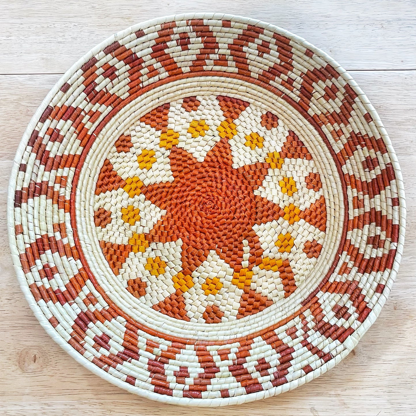 This image features a handcrafted bowl with a detailed pattern woven from natural fibers. At the center is a warm orange floral motif, surrounded by smaller blooms in orange and yellow, all against a beige background. The pattern is framed by concentric circles with geometric designs in earthy tones of beige, brown, and deep red, creating a harmonious and traditional aesthetic.