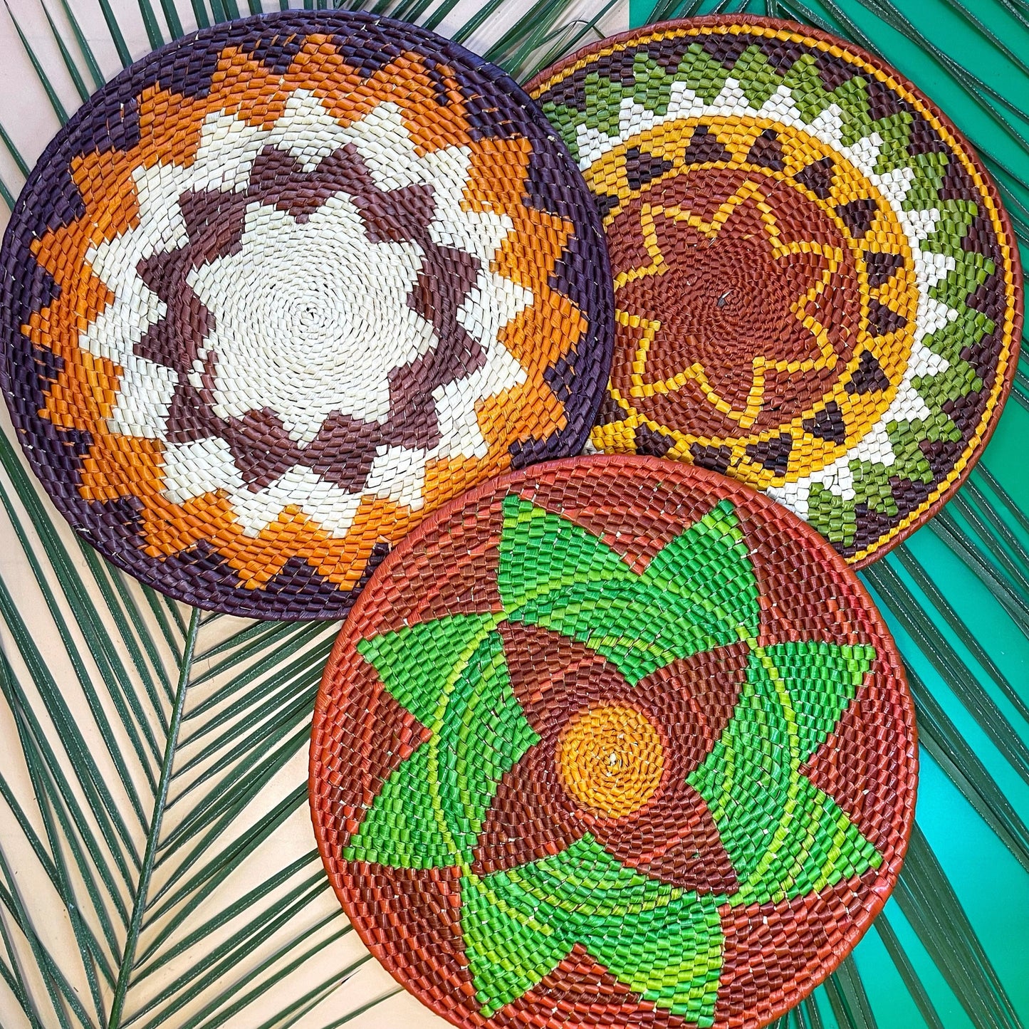 Three circular handwoven placemats made from tucuma palm fibers, displayed on a background with palm fronds. The placemats feature intricate geometric and floral patterns in earthy browns, vivid greens, warm yellows, crisp whites, and rich purples, showcasing traditional craftsmanship.