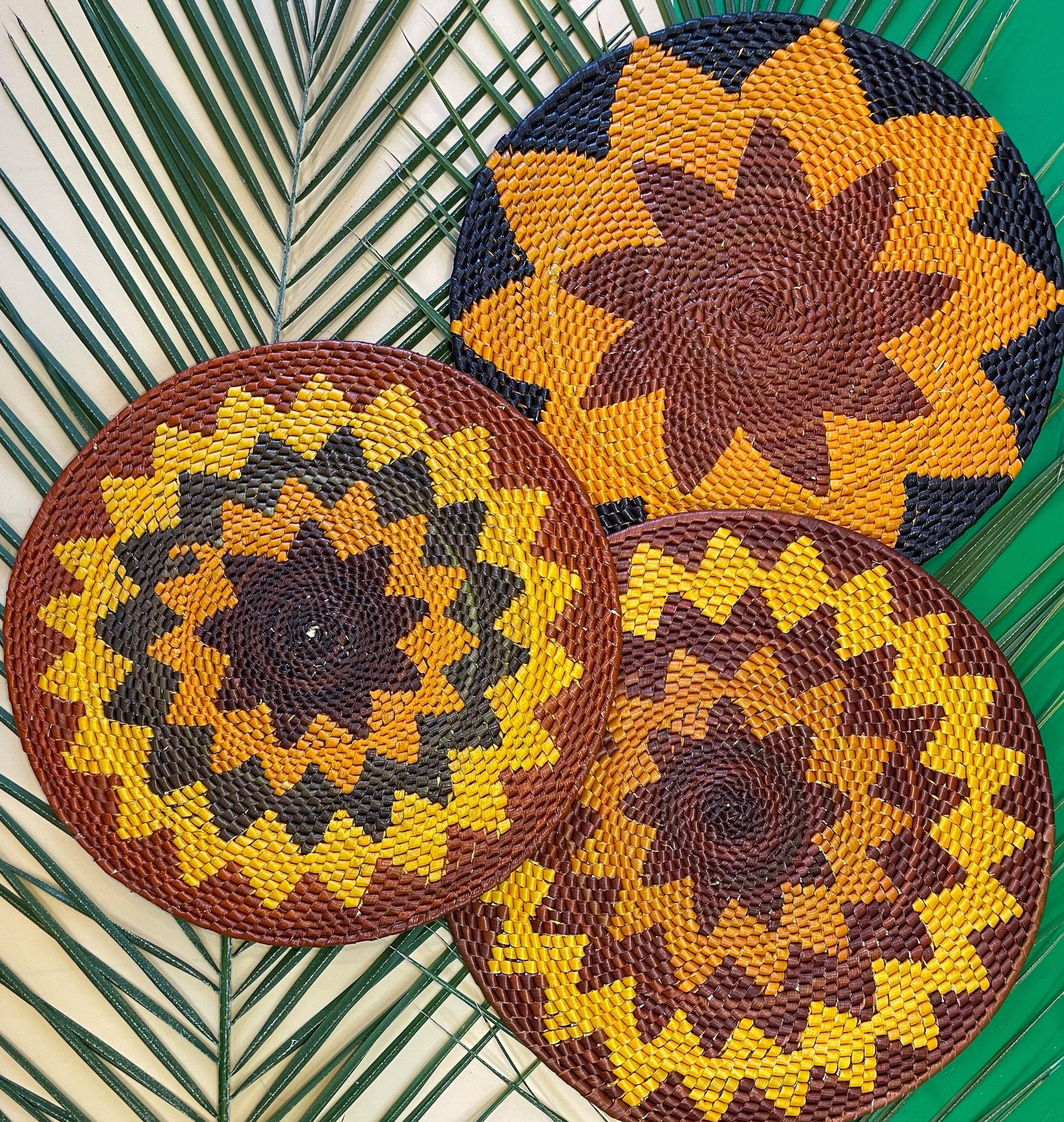 Three handwoven tucuma placemats feature starburst patterns in brown, black, orange, and shades of yellow, laid over palm leaves on a teal background.