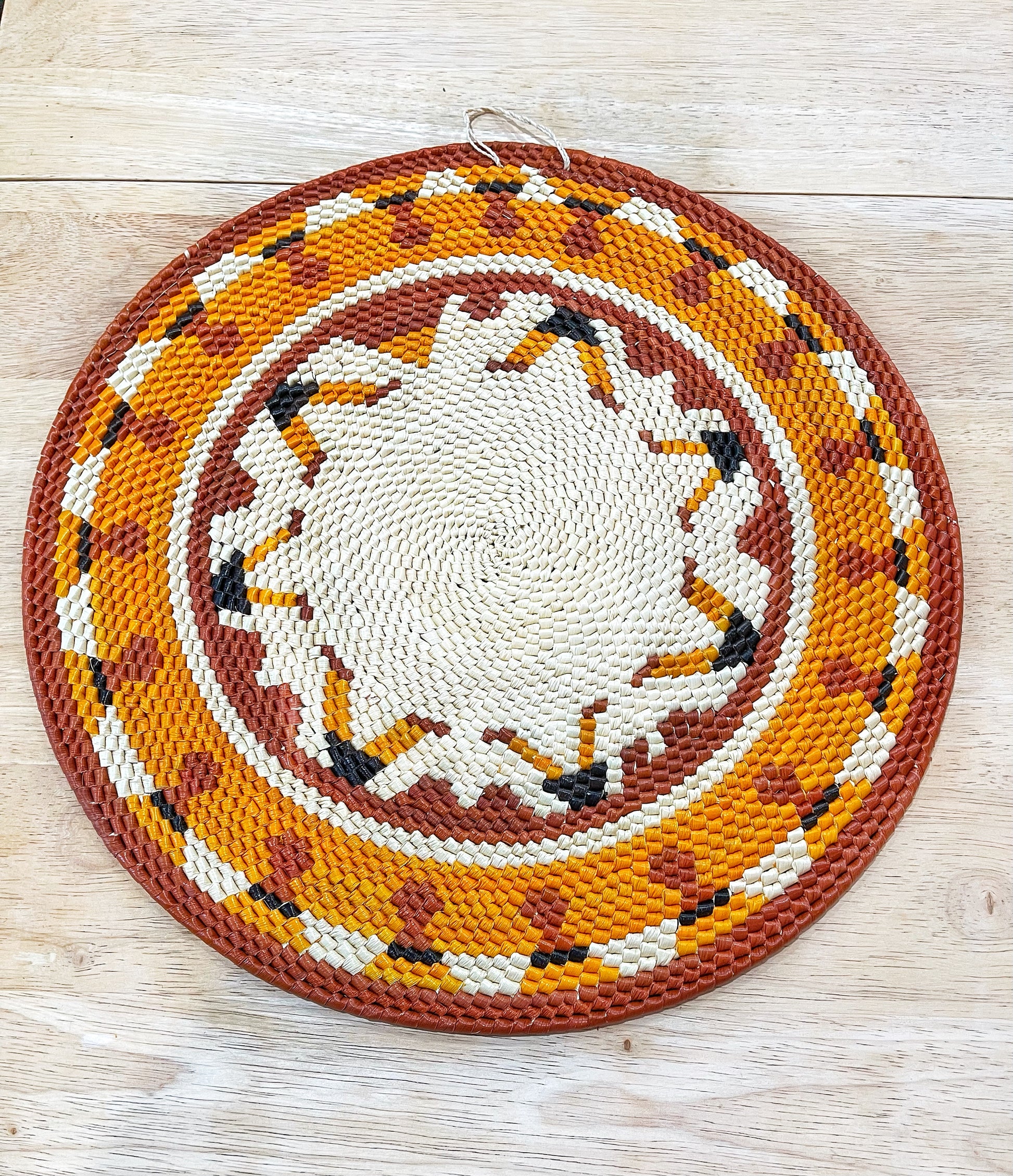 A handwoven mandala wall art in warm hues of orange, rust, cream, and accents of black, featuring a concentric circle pattern with intricate tribal motifs.