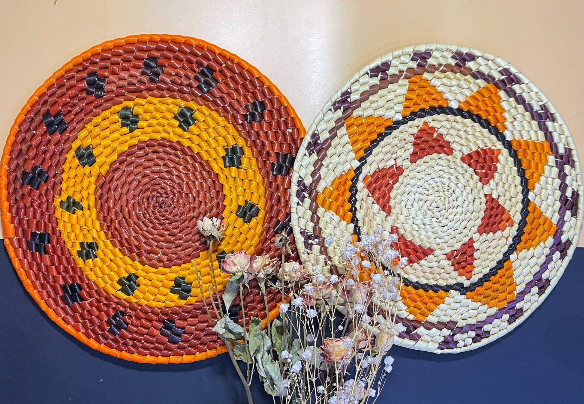 The photo shows two circular handwoven trivets: one with warm shades of red, orange, and accents of dark blue and green; the other features a geometric design with white, orange, red, purple and brown hues, alongside dried flowers.