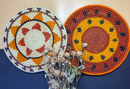 Two handwoven trivets: round shapes with contrasting colors. One features warm red & orange with black accents. The other showcases a geometric pattern in white, orange, red, & purple. Dried flowers nearby.