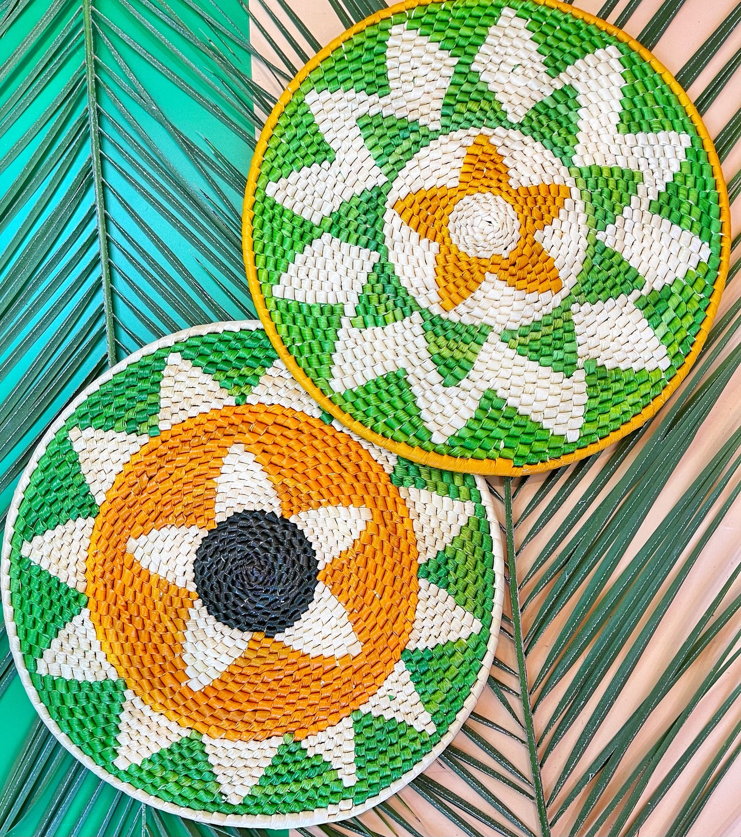 Two round tucuma placemats, one with a green and white spiral, the other with an orange star pattern, on palm leaves.