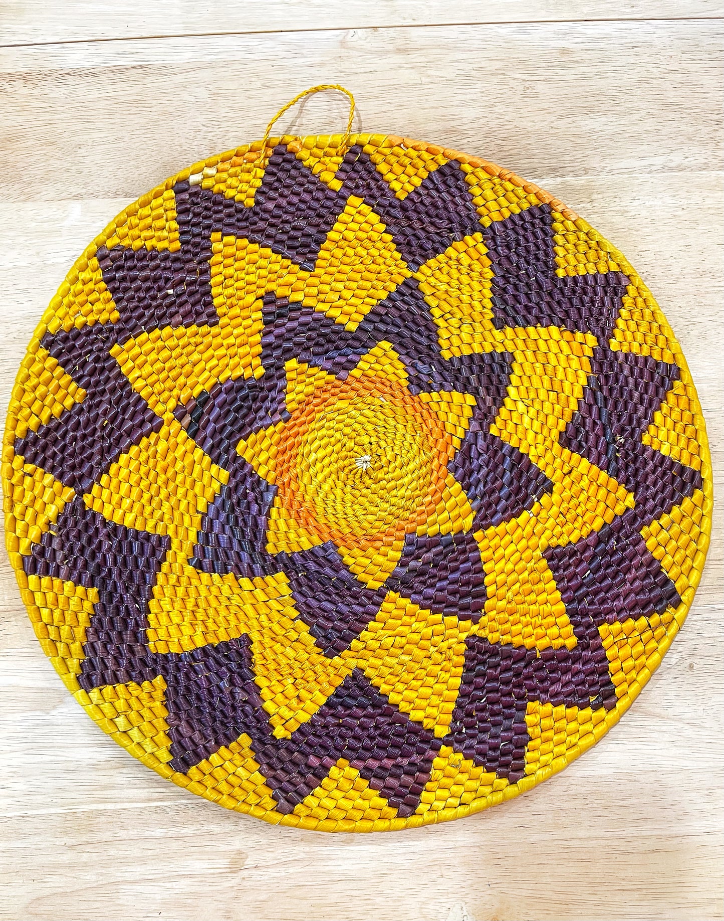 The mandala features a vibrant sunburst pattern with alternating rays of golden yellow and deep purple against a pale yellow center, creating a striking contrast. The woven design adds rich texture.