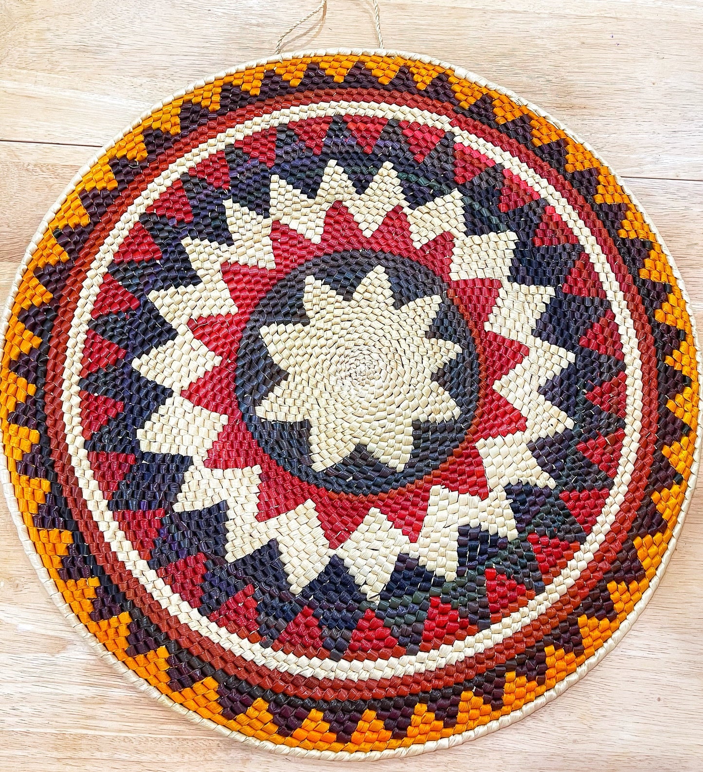 A striking handwoven Amazon mandala wall art piece, featuring a vibrant radial pattern in beige, red, purple, and orange. 