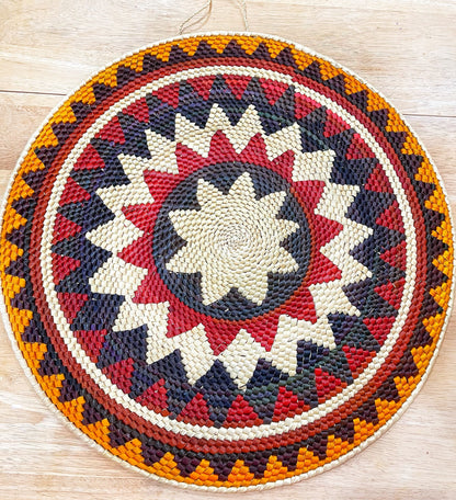 A striking handwoven Amazon mandala wall art piece, featuring a vibrant radial pattern in beige, red, purple, and orange. 