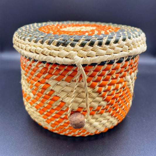 Colorful handwoven tucuma palm box with a tight-fitting lid, featuring vibrant orange, natural beige, and black patterns, and a looped closure with a wooden bead.