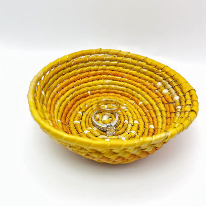 The woven bowl has a beige center, with spiraling rings of yellow and orange. Inside, it holds rings, demonstrating its purpose for small items.