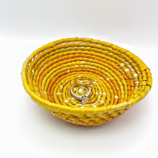 The woven bowl has a beige center, with spiraling rings of yellow and orange. Inside, it holds rings, demonstrating its purpose for small items.