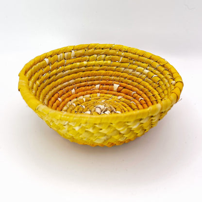 The image displays a handwoven trinket bowl with a natural beige interior that transitions to golden yellow and concludes with a vibrant orange rim. The pattern is coiled and textural.