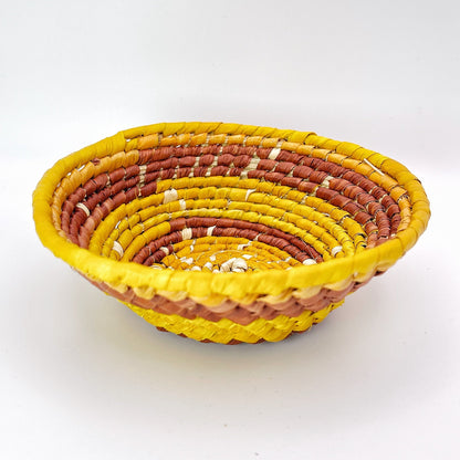 The trinket bowl features concentric circles in beige, transitioning through shades of yellow to an orange-brown, and finally to a vibrant yellow edge, all in a coiled weave.