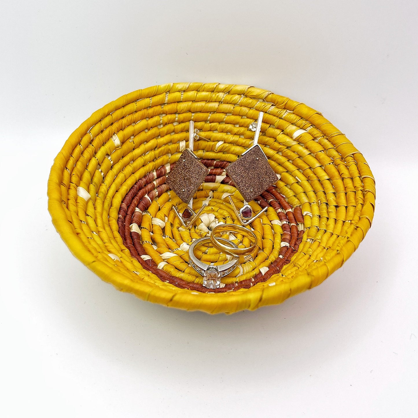 The trinket bowl features a pattern with a light beige center, surrounded by rings of yellow and reddish-brown, and contains earrings and rings, demonstrating its practical use.