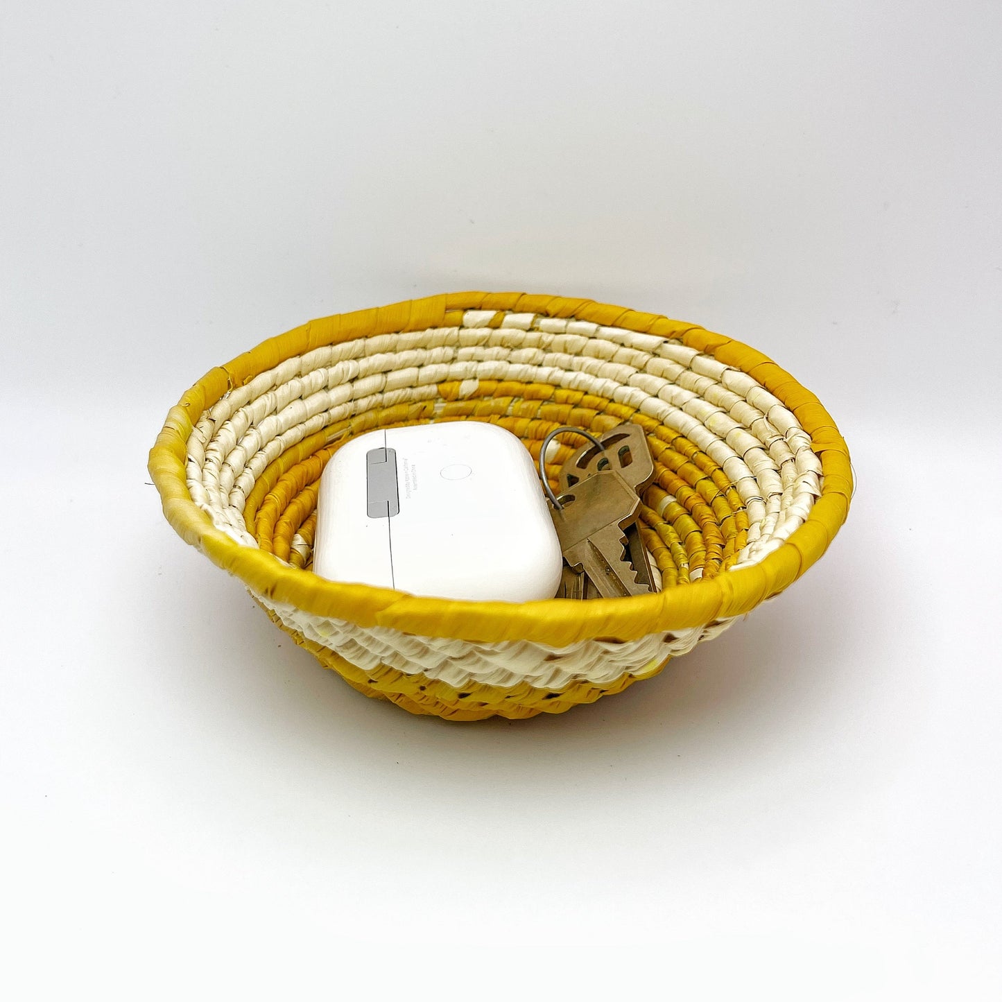 The handwoven trinket bowl features a natural cream interior and a vibrant yellow rim, showcasing the intricate coil pattern of tucuma palm craftsmanship. Inside, an earpod and key demonstrate its storage capacity for small items.