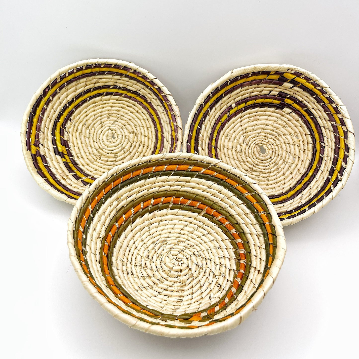 Three trinket bowls feature natural beige, accented with stripes in purple, black, orange, and green, all in a circular pattern emphasizing the handwoven craftsmanship.