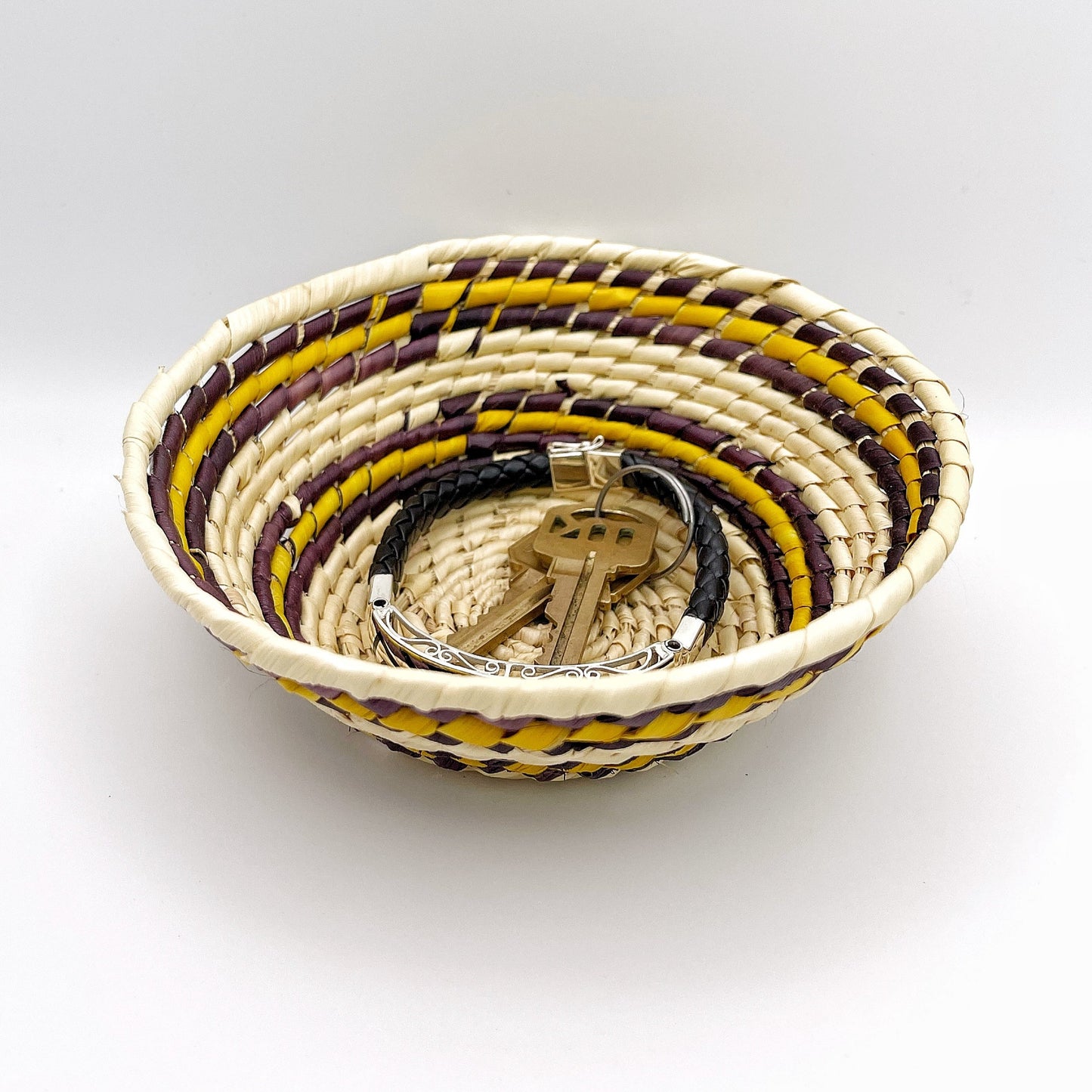 The bowl features a beige base with concentric circles in yellow, brown, and purple. It holds keys and a bracelet, illustrating its use for small items.