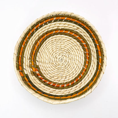 The bowl displays a beige base with concentric rings featuring olive green, orange, and tan, arranged in a pleasing, circular handwoven design.