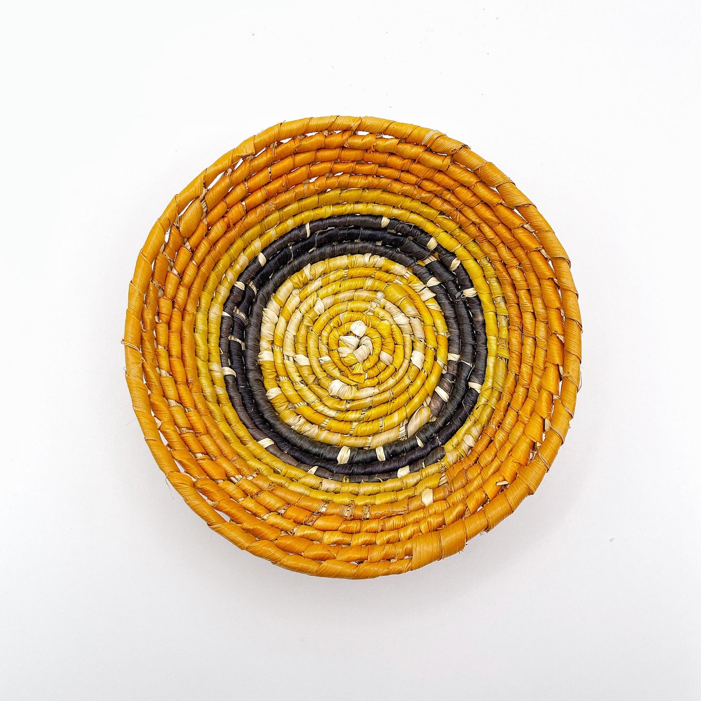 The trinket bowl features a beige center with spirals of yellow, leading out to bands of black and ending in a bold orange edge, highlighting the handcrafted weaving.