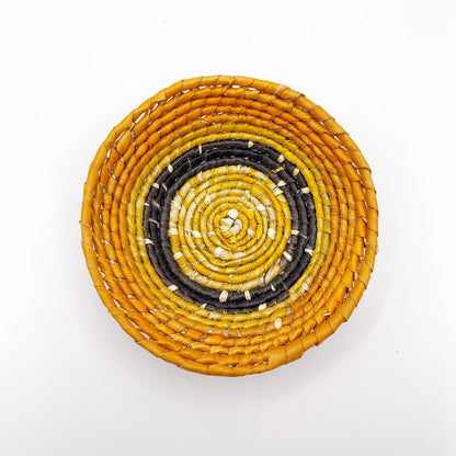 The trinket bowl features a beige center with spirals of yellow, leading out to bands of black and ending in a bold orange edge, highlighting the handcrafted weaving.