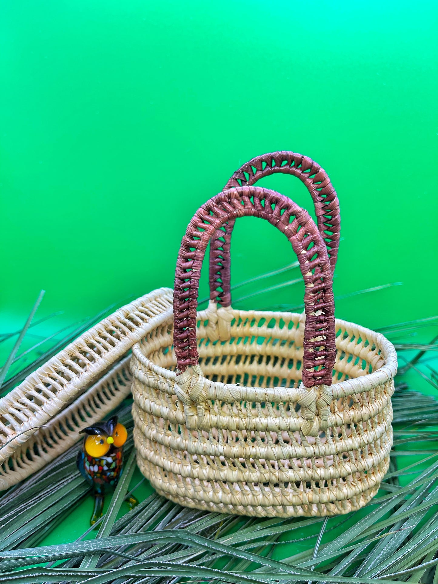 Mini handwoven Amazonian basket: natural beige tucuma fibers, brown accents. Tight weave. Colorful bird figurine beside basket, green backdrop.