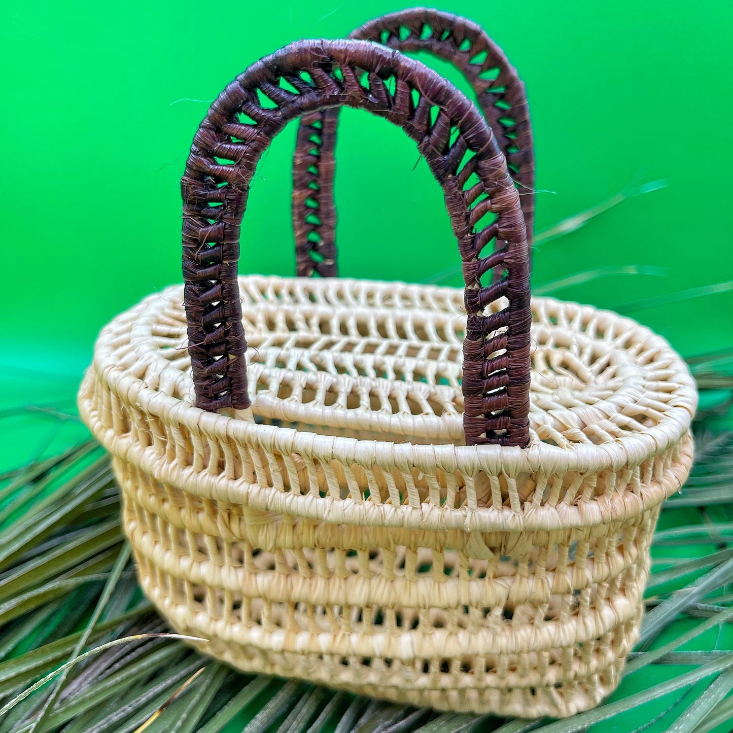  This image features a miniature handwoven basket crafted from tucuma palm fibers. The basket is primarily a natural beige color, showcasing the clean, warm hues of the fibers. It features a sturdy double-handle design, which is wrapped in a dark brown, almost burgundy color, creating a subtle yet attractive contrast. 