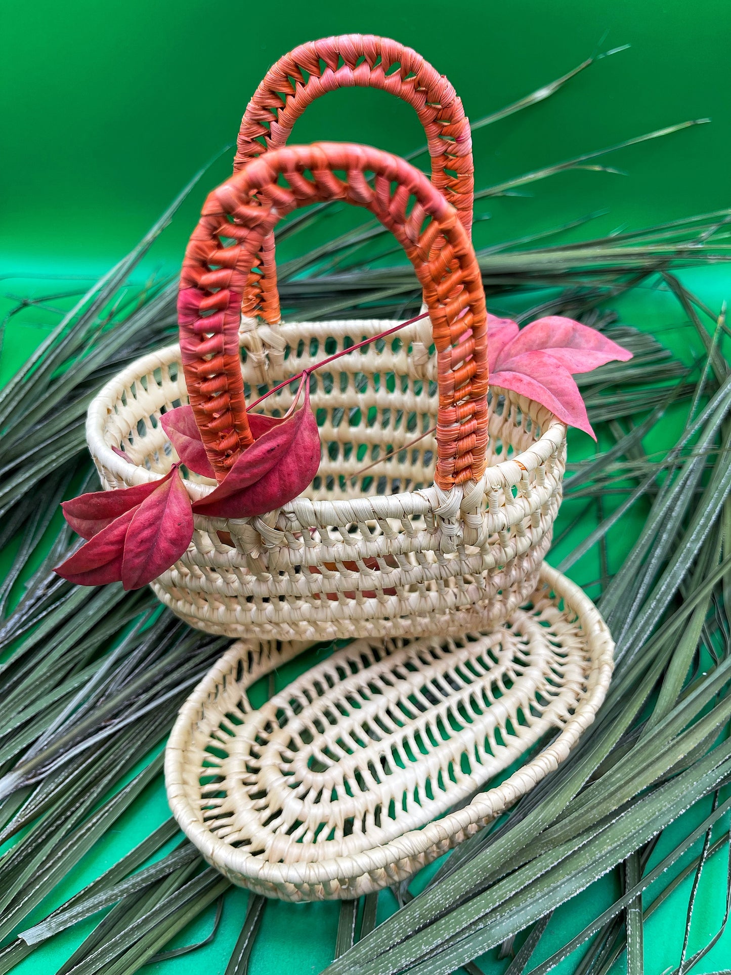  Handwoven mini tucuma basket with vibrant red handles, adorned with pink leaves against a green background with palm sprigs.