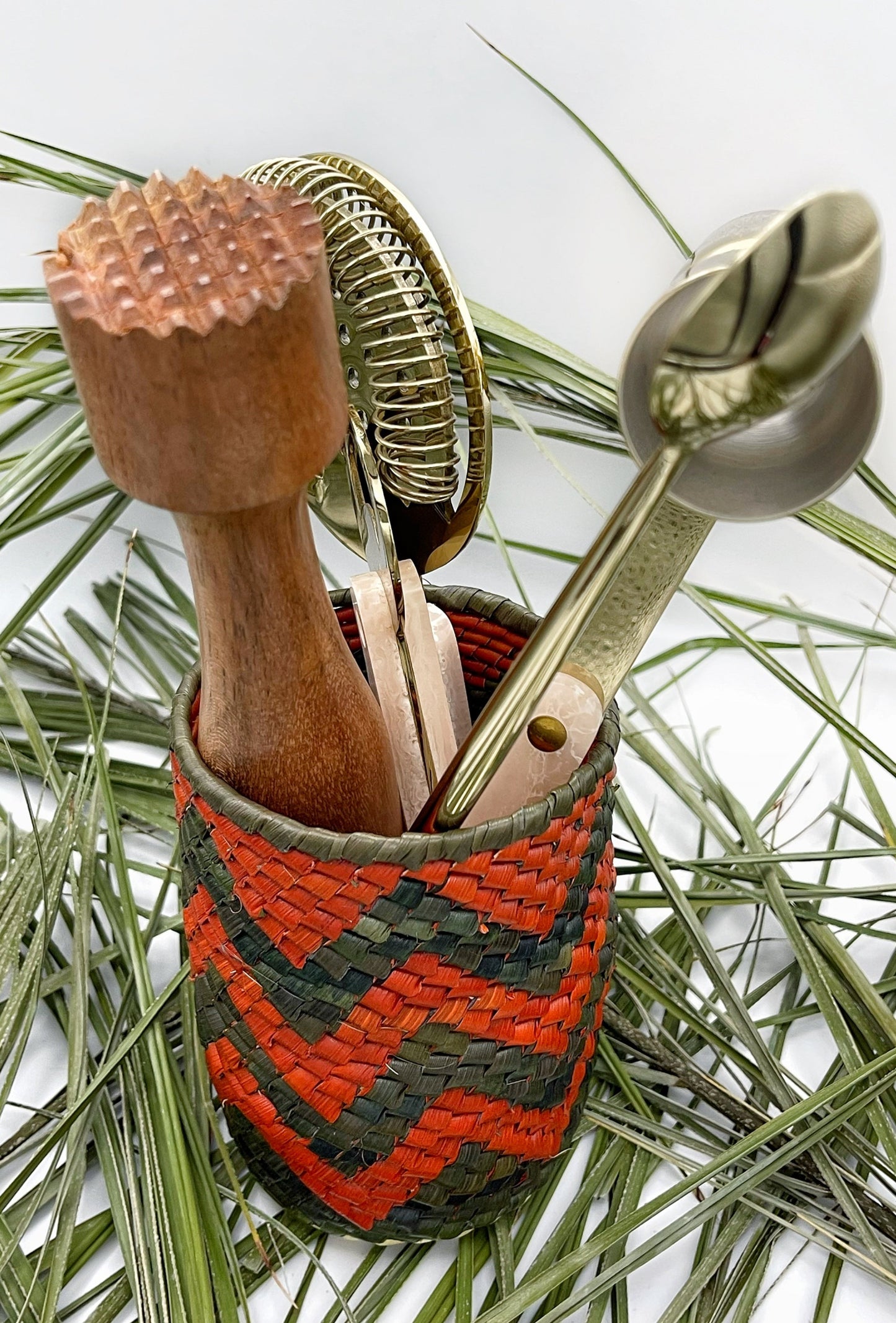 Woven holder with a red and black checkered pattern, holding kitchen tools, set against green pine needles.